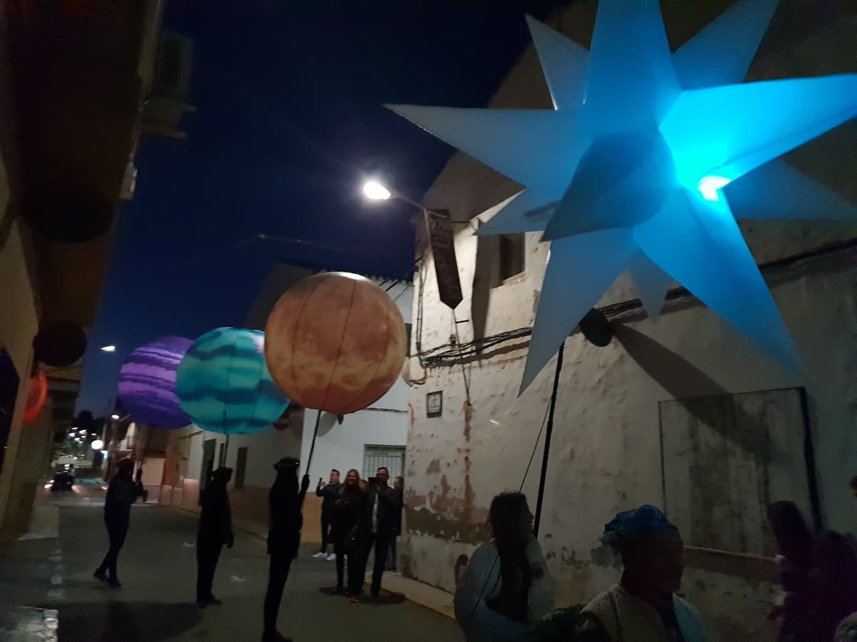 Esferas de planetas y una estrella azul iluminadas junto a una fachada envejecida, con público fotografiando la escena