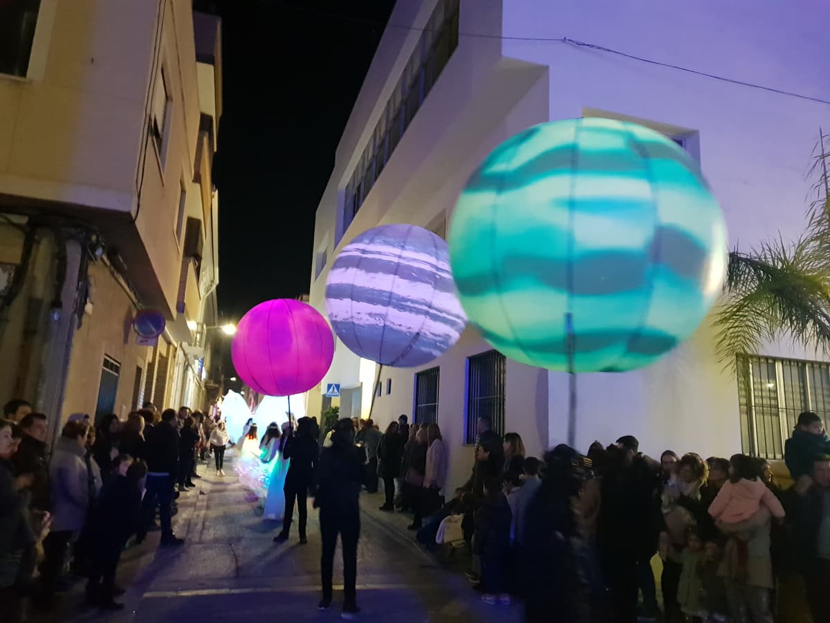 Tres esferas de planeta iluminadas en rosa, morado y turquesa flotando sobre el público en una calle nocturna